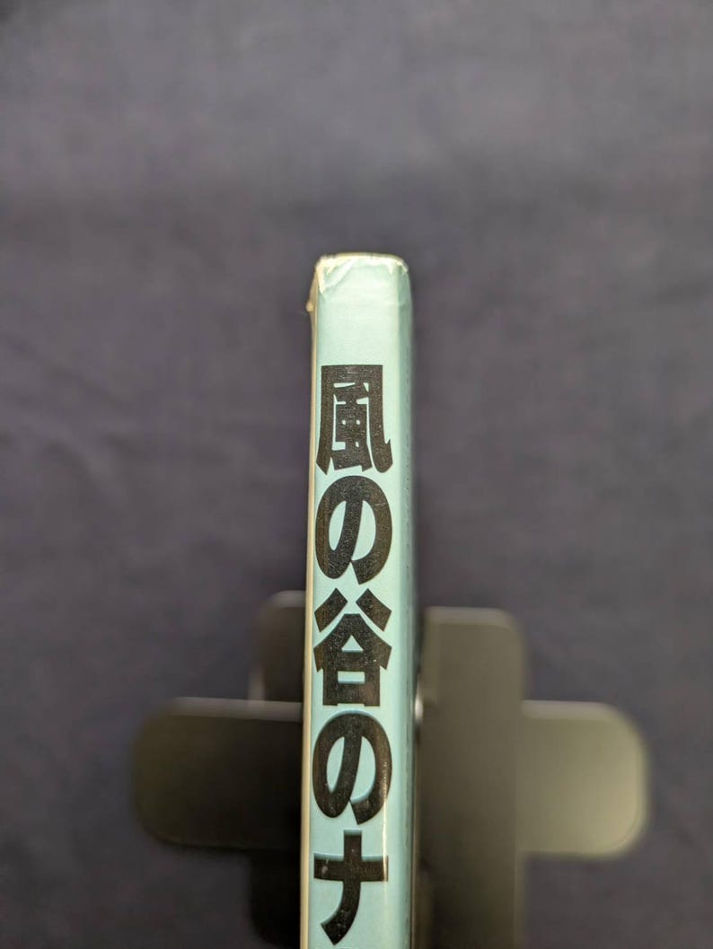 May include: A light blue book spine with Japanese text in black. The book is held in a black book holder against a dark blue background. The text reads "風の谷のナウシカ".
