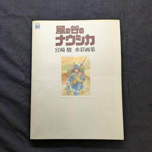 Pu&ograve; includere: Un libro con testo e illustrazioni giapponesi. La copertina presenta un acquerello di un personaggio in abiti blu, che porta un'arma. Il titolo &egrave; in caratteri giapponesi rossi e verdi.