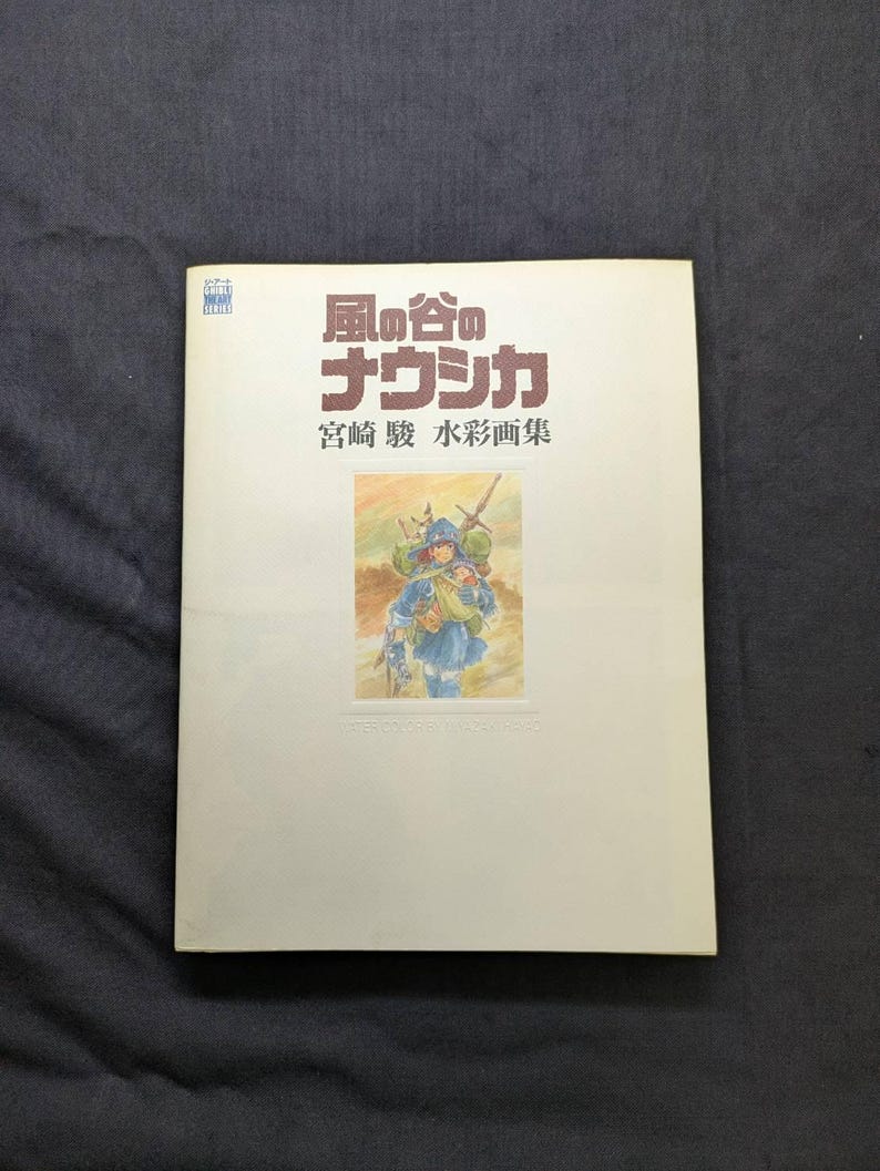 Pu&ograve; includere: Un libro con testo e illustrazioni giapponesi. La copertina presenta un acquerello di un personaggio in abiti blu, che porta un'arma. Il titolo &egrave; in caratteri giapponesi rossi e verdi.