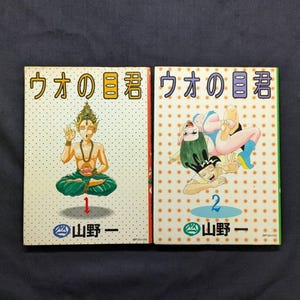 May include: Two Japanese manga books titled "Uo no Me-kun". The first cover features a seated figure with a golden headdress, while the second shows a woman in a dynamic pose. Both books have colorful illustrations and Japanese text.