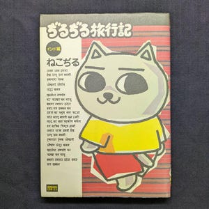 May include: A Japanese comic book cover with a cartoon cat character wearing a yellow shirt and red trousers. The cover features Japanese text and a red and white striped background. The comic book is titled "Zuru Zuru Ryokoki".