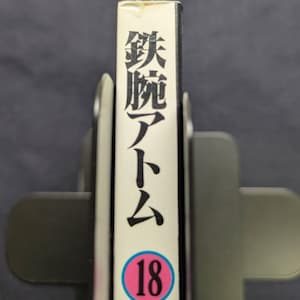 May include: A book spine with Japanese text and the number 18 in a pink circle. The spine is white with black Japanese characters and the title "Astro Boy". The book is held in a gray book holder.