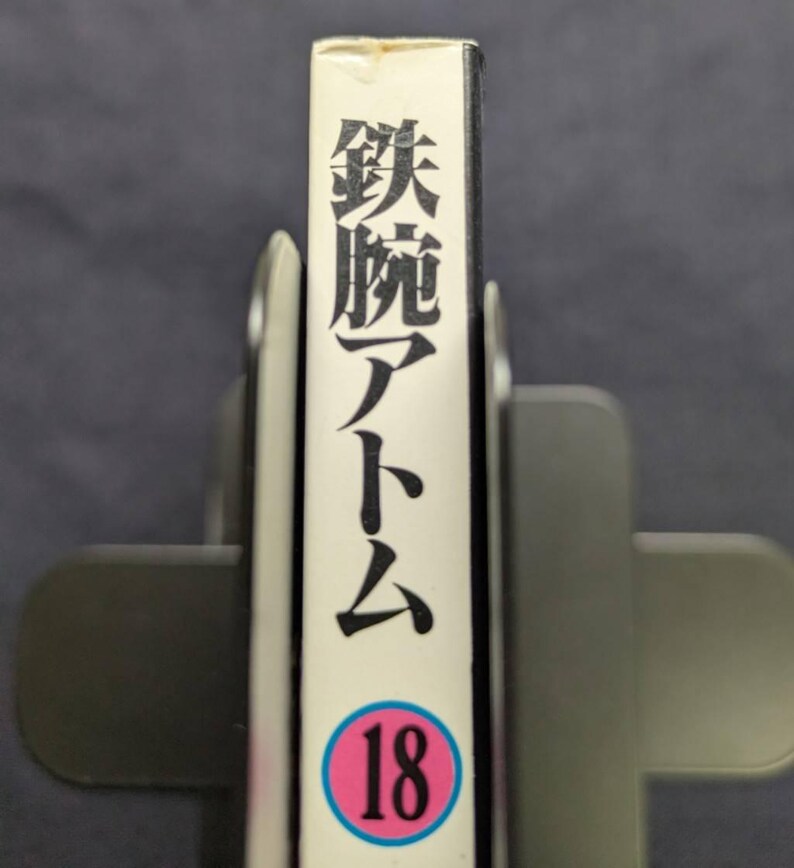 May include: A book spine with Japanese text and the number 18 in a pink circle. The spine is white with black Japanese characters and the title "Astro Boy". The book is held in a gray book holder.