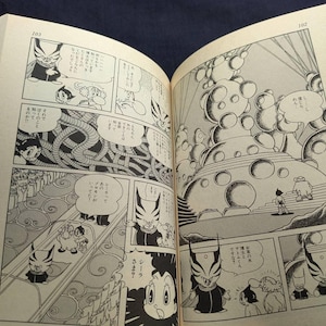 May include: Open book with black and white manga illustrations. The panels depict characters with distinctive features, including a figure with a horned mask and a robot-like character. Text bubbles contain Japanese text.