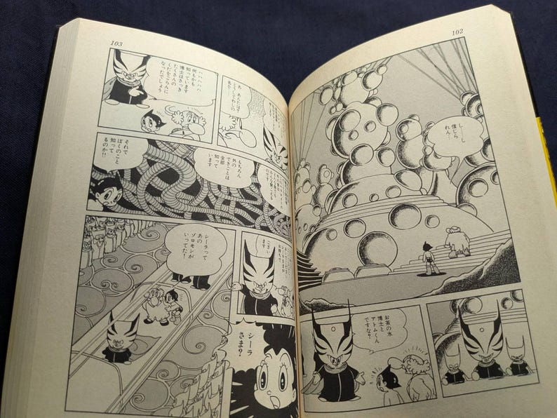 May include: Open book with black and white manga illustrations. The panels depict characters with distinctive features, including a figure with a horned mask and a robot-like character. Text bubbles contain Japanese text.