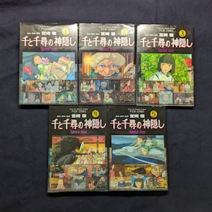 May include: Five Japanese film comics of "Spirited Away" are displayed. Each book has a black cover with Japanese text and a colorful illustration. The books are numbered 1 through 5. The background is a dark blue fabric.