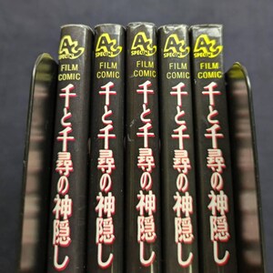 Pode incluir: Uma fila de cinco livros pretos "Film Comic" com texto japon&ecirc;s e o t&iacute;tulo "Spirited Away" em vermelho e branco. Os livros est&atilde;o em um suporte de metal, com o logotipo "A" em amarelo.