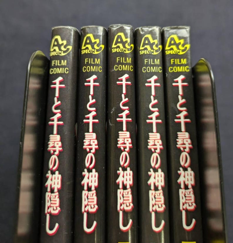 Pode incluir: Uma fila de cinco livros pretos "Film Comic" com texto japon&ecirc;s e o t&iacute;tulo "Spirited Away" em vermelho e branco. Os livros est&atilde;o em um suporte de metal, com o logotipo "A" em amarelo.