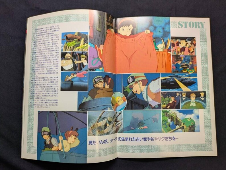 May include: An open book featuring colorful anime-style illustrations from a film story. The pages display multiple scenes, including characters in flight, a person wearing oversized clothing, and text in Japanese. The layout includes the word STORY.