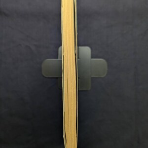 May include: A book with yellowed pages, held upright in a black book stand. The spine is not visible, showing the aged edges of the pages. The book is set against a dark blue background.