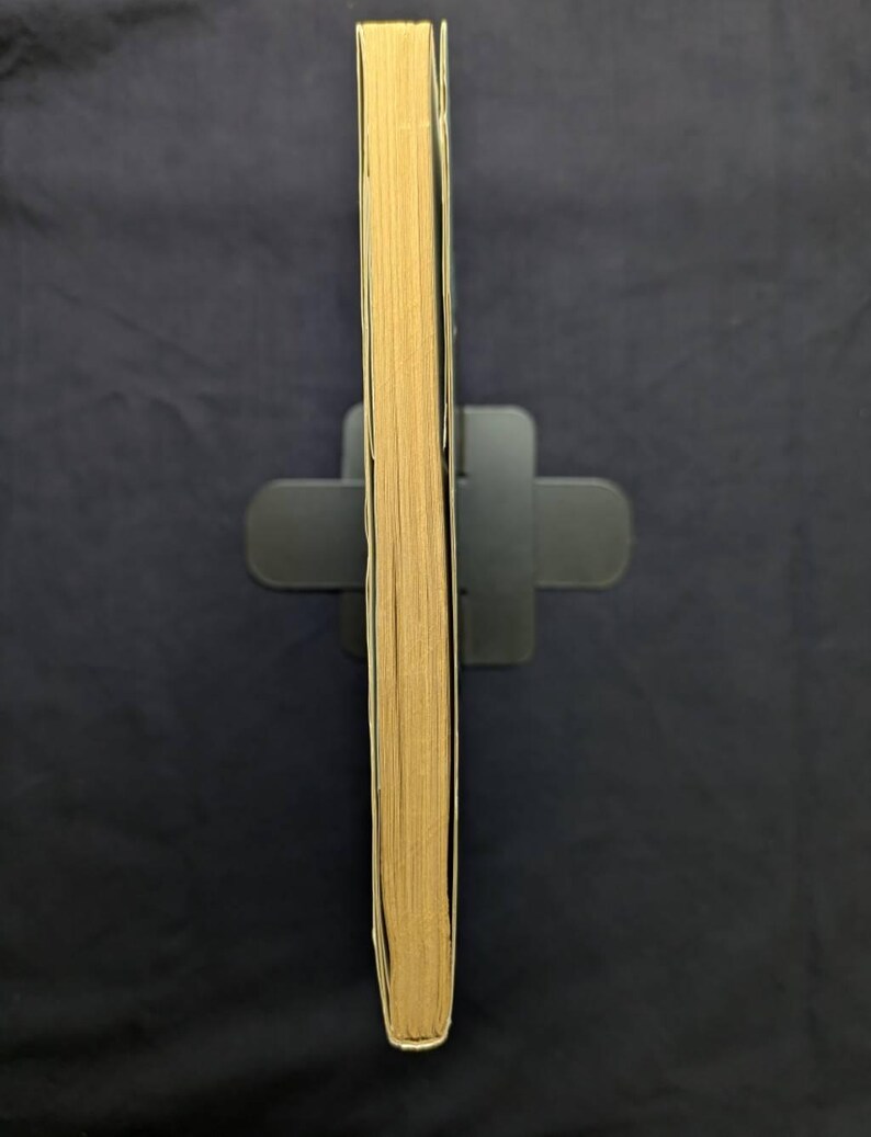 May include: A book with yellowed pages, held upright in a black book stand. The spine is not visible, showing the aged edges of the pages. The book is set against a dark blue background.