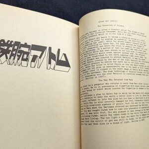 May include: Open book with text and a large, stylized Japanese text in black. The text is on a cream-colored page. The book is open to a page with a title and more text.
