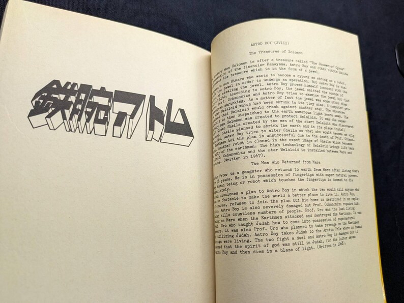 May include: Open book with text and a large, stylized Japanese text in black. The text is on a cream-colored page. The book is open to a page with a title and more text.