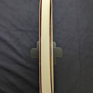 Pu&ograve; includere: Un libro spesso con pagine color crema e dorso rosso, tenuto in posizione verticale da un leggio grigio. I bordi del libro sono visibili, mostrando le pagine impilate. Lo sfondo &egrave; un tessuto grigio scuro.