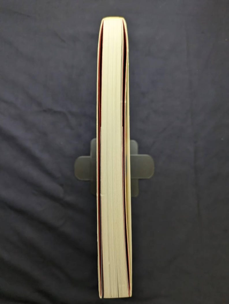 Pu&ograve; includere: Un libro spesso con pagine color crema e dorso rosso, tenuto in posizione verticale da un leggio grigio. I bordi del libro sono visibili, mostrando le pagine impilate. Lo sfondo &egrave; un tessuto grigio scuro.