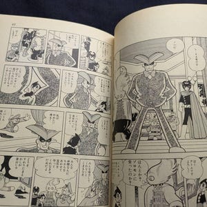 May include: Open book with Japanese manga illustrations. The black and white panels depict various characters in action, with speech bubbles containing Japanese text. The pages are yellowed with age, suggesting a vintage comic book.
