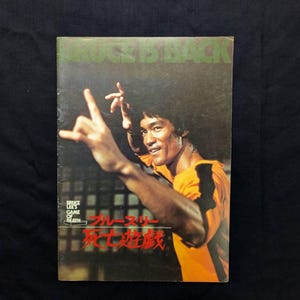 以下が含まれることがあります： 黄色と黒のジャンプスーツを着て武道のポーズで腕を上げたブルース・リーが写る長方形の映画パンフレット。上部に「BRUCE IS BACK」、下部に「BRUCE LEE'S GAME OF DEATH」の文字と日本語のタイトルが記載されています。