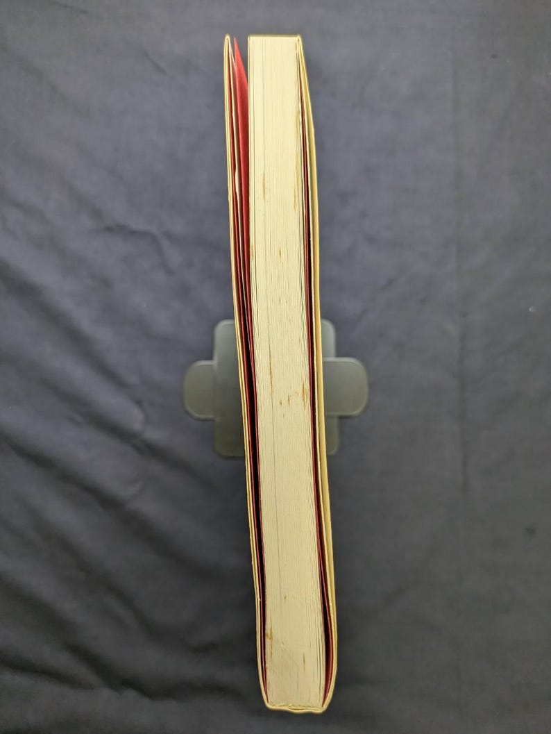 Pu&ograve; includere: Un libro con copertina rigida con una copertina color crema e bordi delle pagine rossi. Il libro &egrave; tenuto in posizione verticale da un leggio grigio. Le pagine sono visibili e il dorso &egrave; leggermente usurato. Lo sfondo &egrave; un tessuto grigio scuro.