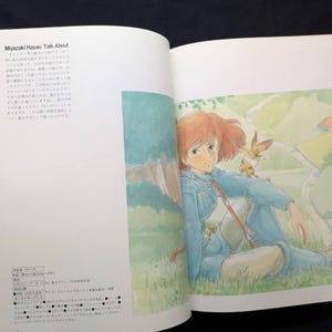 Pu&ograve; includere: Libro aperto con un'illustrazione colorata di una persona con i capelli rossi, vestita di blu, seduta in un'area erbosa. Un piccolo uccello &egrave; posato sulla sua spalla. Il testo sulla pagina di sinistra recita "Miyazaki Hayao Talk About".
