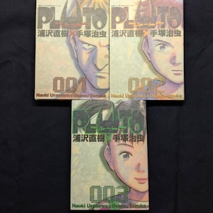 May include: Three manga volumes of "Pluto" by Naoki Urasawa and Osamu Tezuka are displayed. Each volume features a different character portrait and the title in Japanese. The volumes are numbered 001, 002, and 003.