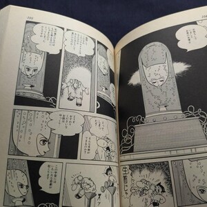 May include: Open book with black and white manga illustrations. Panels depict a character in a glass container, with speech bubbles containing Japanese text. The art style is vintage, with detailed linework and shading.