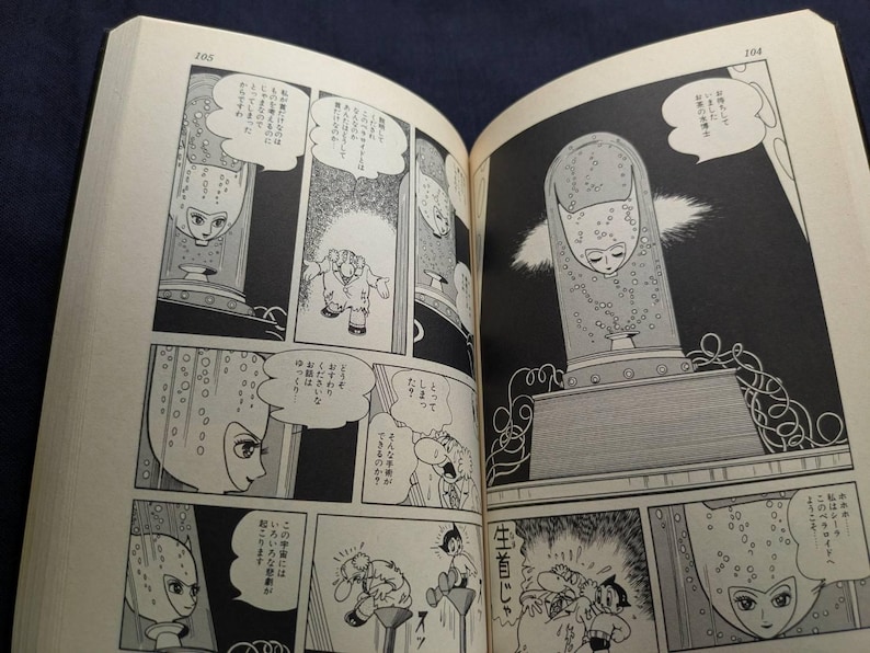 May include: Open book with black and white manga illustrations. Panels depict a character in a glass container, with speech bubbles containing Japanese text. The art style is vintage, with detailed linework and shading.