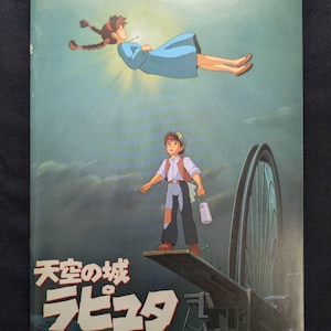 May include: A rectangular poster featuring an animated scene with a person in a blue dress floating in the sky above a person standing on a wooden plank. The text "天空の城ラピュタ" is printed in large white characters at the bottom left corner.