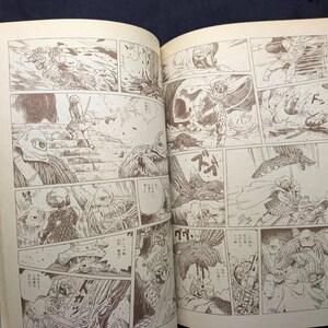 May include: Open book with manga-style illustrations in sepia tones. The panels depict action scenes with figures in helmets and armor, set in a fantasy or science fiction world. The pages are off-white with a light blue spine.