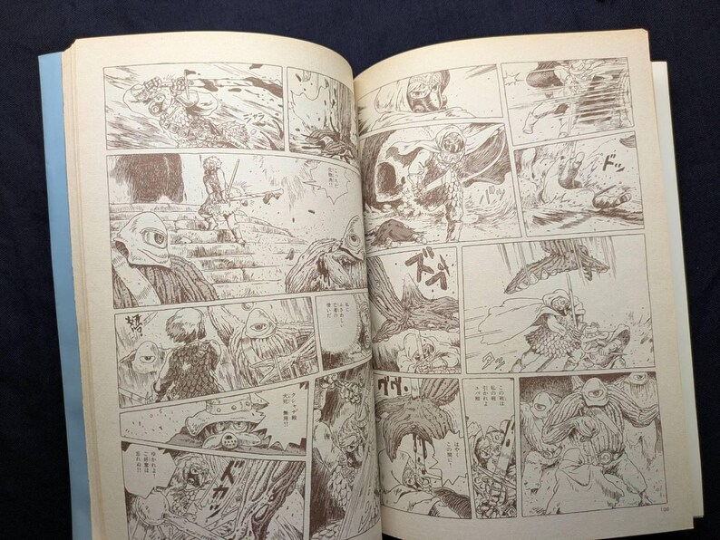 May include: Open book with manga-style illustrations in sepia tones. The panels depict action scenes with figures in helmets and armor, set in a fantasy or science fiction world. The pages are off-white with a light blue spine.