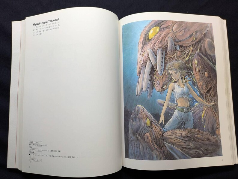 Pu&ograve; includere: Libro aperto con un'illustrazione colorata di una giovane donna con top bianco e pantaloni blu, in piedi vicino a una grande creatura marrone simile a un mostro. L'illustrazione &egrave; in uno stile anime dettagliato. Il testo sulla pagina di sinistra recita "Miyazaki Hayao Talk About".
