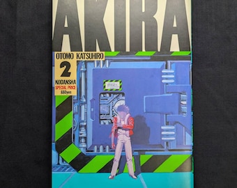 May include: A copy of the manga "AKIRA" volume 2. The cover features the title in large white block letters, the author's name, and a futuristic illustration of a character in a red jacket. The background is blue and green.