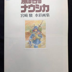 Pu&ograve; includere: Un libro bianco con testo giapponese e un'illustrazione ad acquerello. Il titolo "Nausica&auml; della Valle del Vento" &egrave; visualizzato in grandi caratteri marroni. L'illustrazione raffigura una figura in abiti blu.
