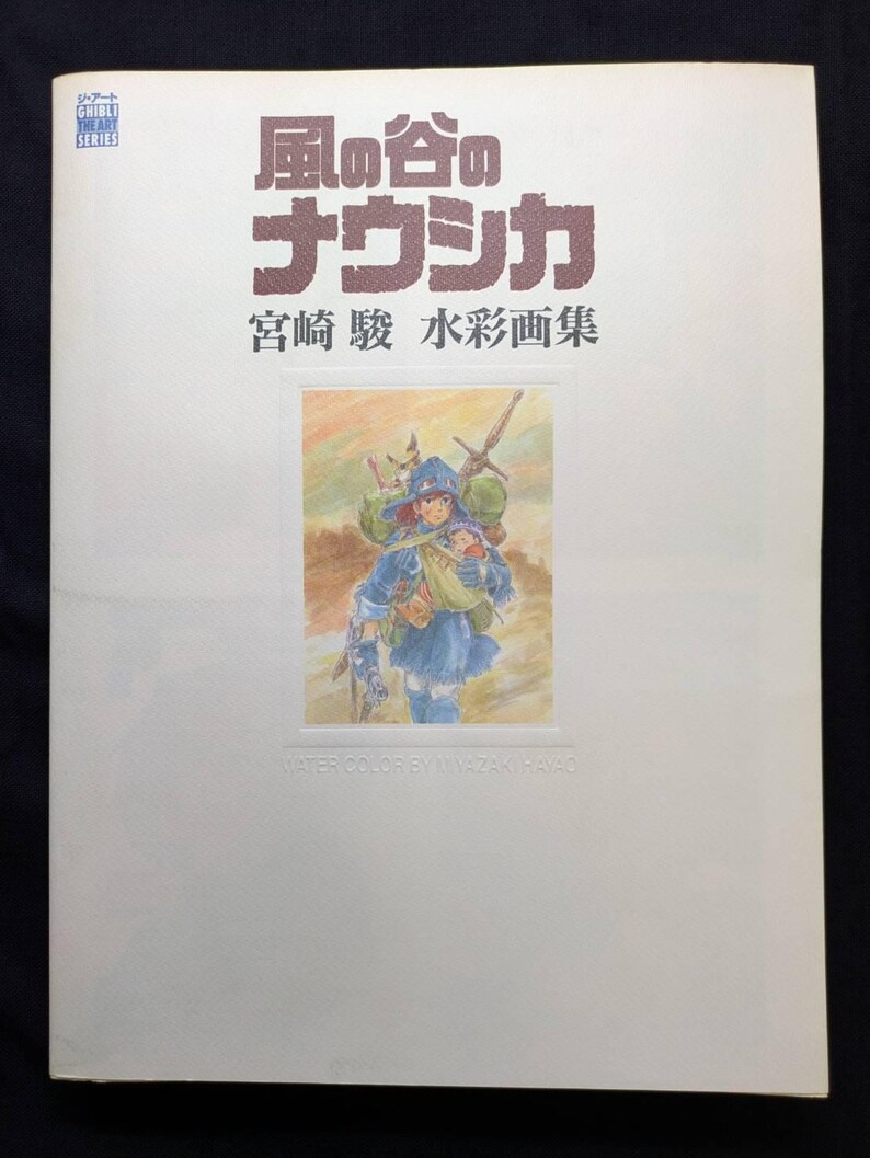 Pu&ograve; includere: Un libro bianco con testo giapponese e un'illustrazione ad acquerello. Il titolo "Nausica&auml; della Valle del Vento" &egrave; visualizzato in grandi caratteri marroni. L'illustrazione raffigura una figura in abiti blu.