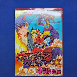 May include: A rectangular manga book cover featuring colorful anime-style characters, including a figure in a red hood holding a grey handgun. The Japanese text reads ヴァンパイア セイヴァー 4 決定版. The background displays vibrant orange and yellow flames against a blue sky.