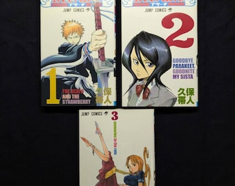 May include: Three manga volumes of the series "Bleach" are displayed. The covers feature colorful illustrations of characters and the series title. Volume 1 is titled "The Death and the Strawberry". Volume 2 is titled "Goodbye Parakeet, Goodnite My Sista". Volume 3 is titled "Memories in the Rain".