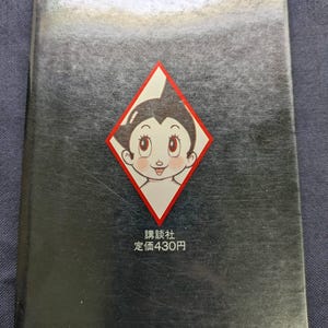 May include: A vintage book with a black cover featuring a diamond-shaped graphic of a cartoon character with large eyes. The text "講談社 定価430円" is printed below the graphic. The book's spine has additional text.