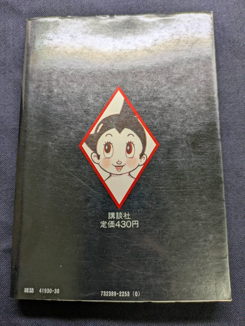 May include: A vintage book with a black cover featuring a diamond-shaped graphic of a cartoon character with large eyes. The text "講談社 定価430円" is printed below the graphic. The book's spine has additional text.