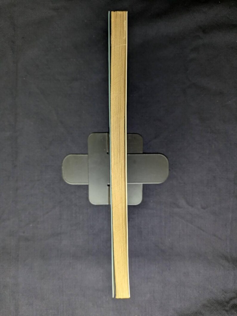 May include: A stack of book pages with a light tan color, held upright by a black book stand. The pages are slightly angled, revealing the edges. The background is a dark blue fabric.
