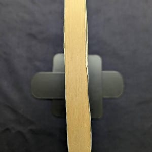 May include: A light brown book with a white border is held upright by a black book stand. The book's pages are visible, and the spine is facing the viewer. The background is a dark, solid color.