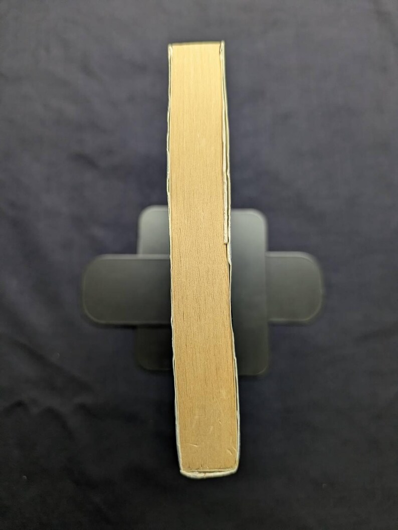 May include: A light brown book with a white border is held upright by a black book stand. The book's pages are visible, and the spine is facing the viewer. The background is a dark, solid color.