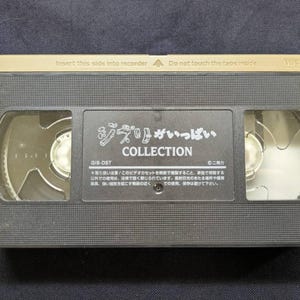 Op de afbeelding: Een zwarte VHS-videoband met gouden details. Het label bevat Japanse tekst, waaronder "ジブリがいっぱい COLLECTION". De bovenkant van de band leest "Plaats deze kant in de recorder" en "Raak de tape binnenin niet aan".