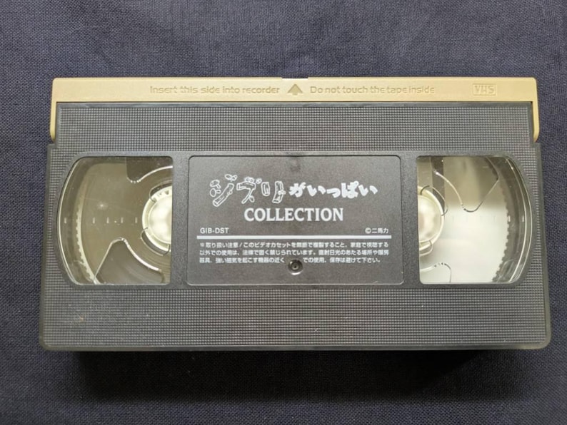 Op de afbeelding: Een zwarte VHS-videoband met gouden details. Het label bevat Japanse tekst, waaronder "ジブリがいっぱい COLLECTION". De bovenkant van de band leest "Plaats deze kant in de recorder" en "Raak de tape binnenin niet aan".