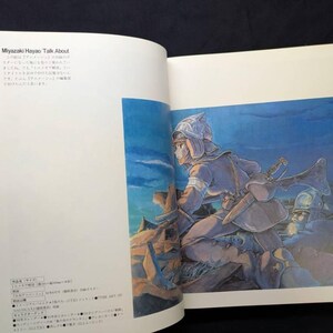 Pu&ograve; includere: Libro aperto con un'illustrazione a tutta pagina di un personaggio in armatura, con una spada. L'opera d'arte &egrave; nei toni del blu, marrone e beige. Testo giapponese visibile sulla pagina sinistra.