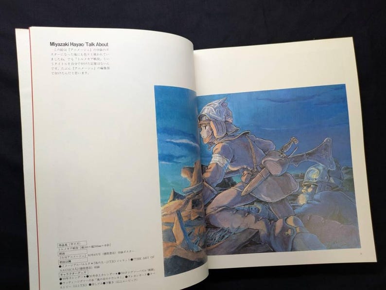 Pu&ograve; includere: Libro aperto con un'illustrazione a tutta pagina di un personaggio in armatura, con una spada. L'opera d'arte &egrave; nei toni del blu, marrone e beige. Testo giapponese visibile sulla pagina sinistra.