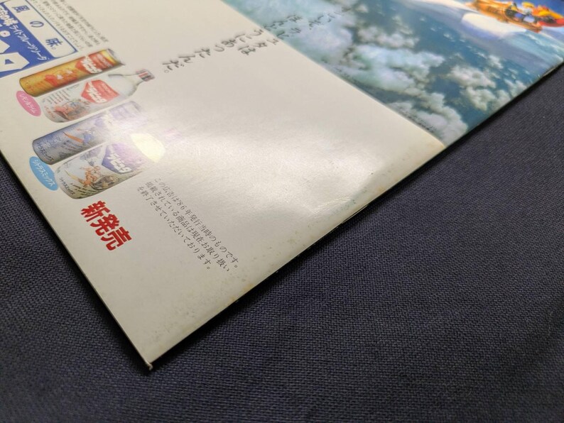 May include: A glossy paper advertisement featuring a blue sky with clouds and a small orange vehicle in the upper right corner. The left side displays four beverage bottles with Japanese text, including the characters 風の味 and 新発売.