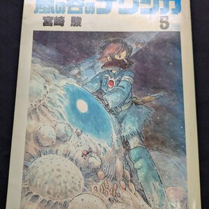 May include: A vintage Japanese manga comic book titled "Nausica&auml; of the Valley of the Wind" with a cover illustration of a character in a blue outfit. The cover features Japanese text and the number 5.