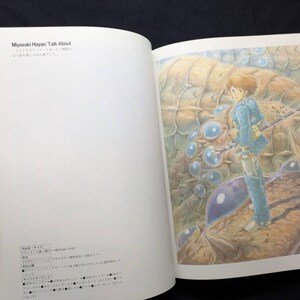 Pu&ograve; includere: Un libro aperto con un'illustrazione a tutta pagina. L'illustrazione raffigura una persona in abito blu che tiene un'arma, in piedi di fronte a una grande creatura strutturata, marrone e blu. Il testo "Miyazaki Hayao Talk About" &egrave; visibile.