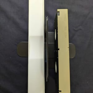 Op de afbeelding: Een witte VHS-bandhoes en een beige VHS-band zijn verticaal geplaatst op een donkere ondergrond. De bandhoes is rechthoekig en de VHS-band heeft labels en een zwart interieur. De items zijn in een zwarte houder geplaatst.