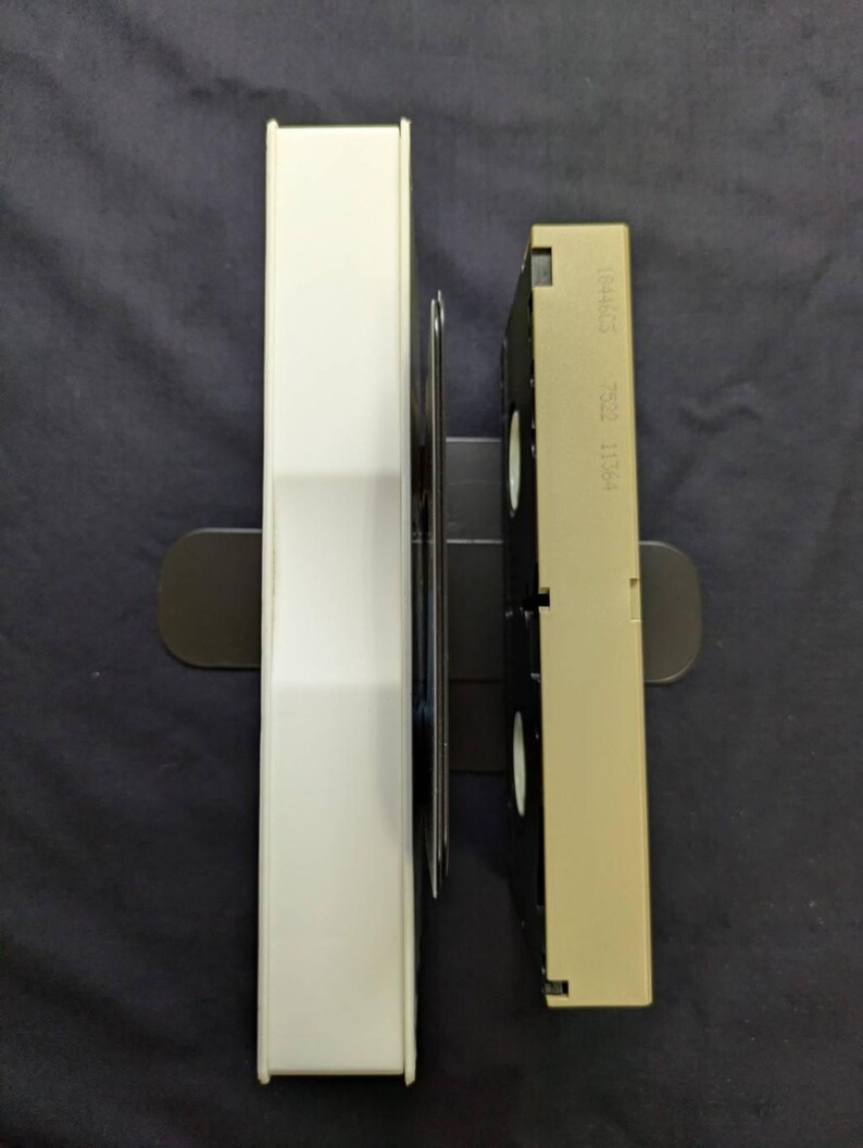 Op de afbeelding: Een witte VHS-bandhoes en een beige VHS-band zijn verticaal geplaatst op een donkere ondergrond. De bandhoes is rechthoekig en de VHS-band heeft labels en een zwart interieur. De items zijn in een zwarte houder geplaatst.
