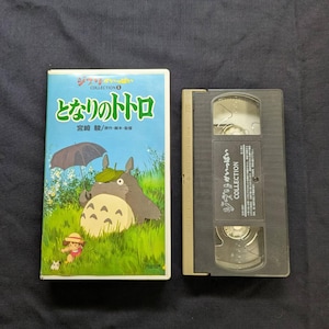 Op de afbeelding: Een VHS-band en hoes met de animatiefilm "My Neighbor Totoro". De hoes is lichtblauw met Japanse tekst en illustraties van Totoro en een jong meisje. De VHS-band is zwart en heeft Japanse tekst.